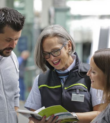 Eine BLS-Reisebegleiterin berät einen Kunden und eine Kundin mit einem BLS-Prospekt in den Händen